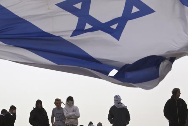 An Israeli flag flutters near Israeli youths as they protest atop the roof of a synagogue in the Israeli settlement of Givat Ze'ev (photo credit: REUTERS)